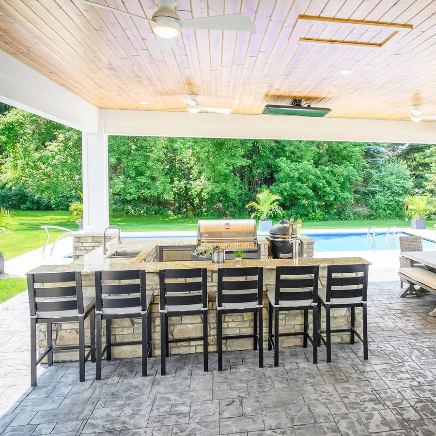 Covered outdoor kitchen with stone bar, built-in grill and bar stools beside a pool